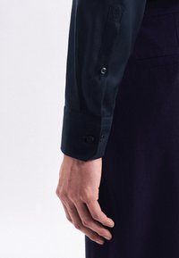 Camicia a maniche lunghe blu navy con tessuto testurizzato, caratterizzata da un polsino con bottoni e un design aderente su misura. Le rifiniture includono un polsino a scallop.