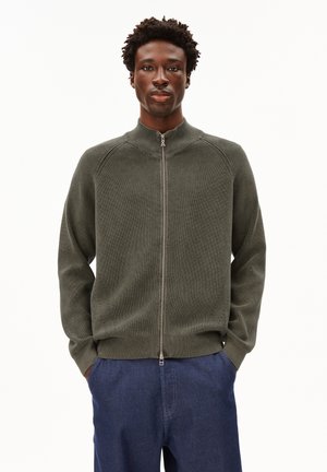 Donkergroene gebreide trui met rits en hoge kraag, met een geribbelde textuur en een taille-lange stijl, gecombineerd met blauwe spijkerbroeken.