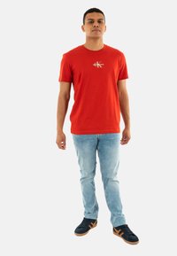 Camiseta de algodón roja con cuello redondo y mangas cortas, que presenta un pequeño logo metálico en el pecho, combinada con jeans de lavado claro y zapatillas negras.