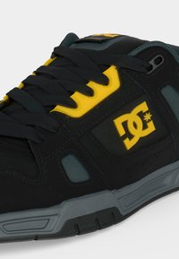 Μαύρο και κίτρινο sneaker μάρκας DC με μαύρα κορδόνια, γκρι σόλα και λεπτομέρειες από δίχτυ στο πάνω μέρος.