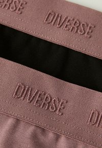 Różowy elastyczny pasek w talii z haftem "Diverse" w ciemniejszym różowym, połączony z czarnym materiałem poniżej. Gładka tekstura i staranne detale szycia.