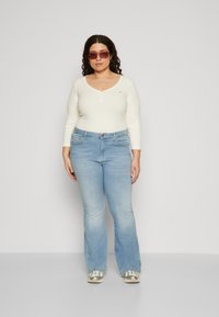 Helle blaue Schlagjeans und ein geripptes cremfarbenes Oberteil mit V-Ausschnitt, das ein kleines Logo trägt. Kombiniert mit übergroßen rosa Sonnenbrillen.
