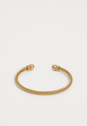 Narukvica od zlatnog toniranog metala s uvijenim dizajnom, glatkim okruglim završecima i teksturiranim detaljima na zatvaranju. Elegantnog i minimalističkog oblika.