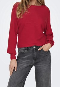 Pull rouge côtelé avec une coupe décontractée et des manches longues, associé à un jean gris foncé. Le pull présente un col large et un tissu texturé.