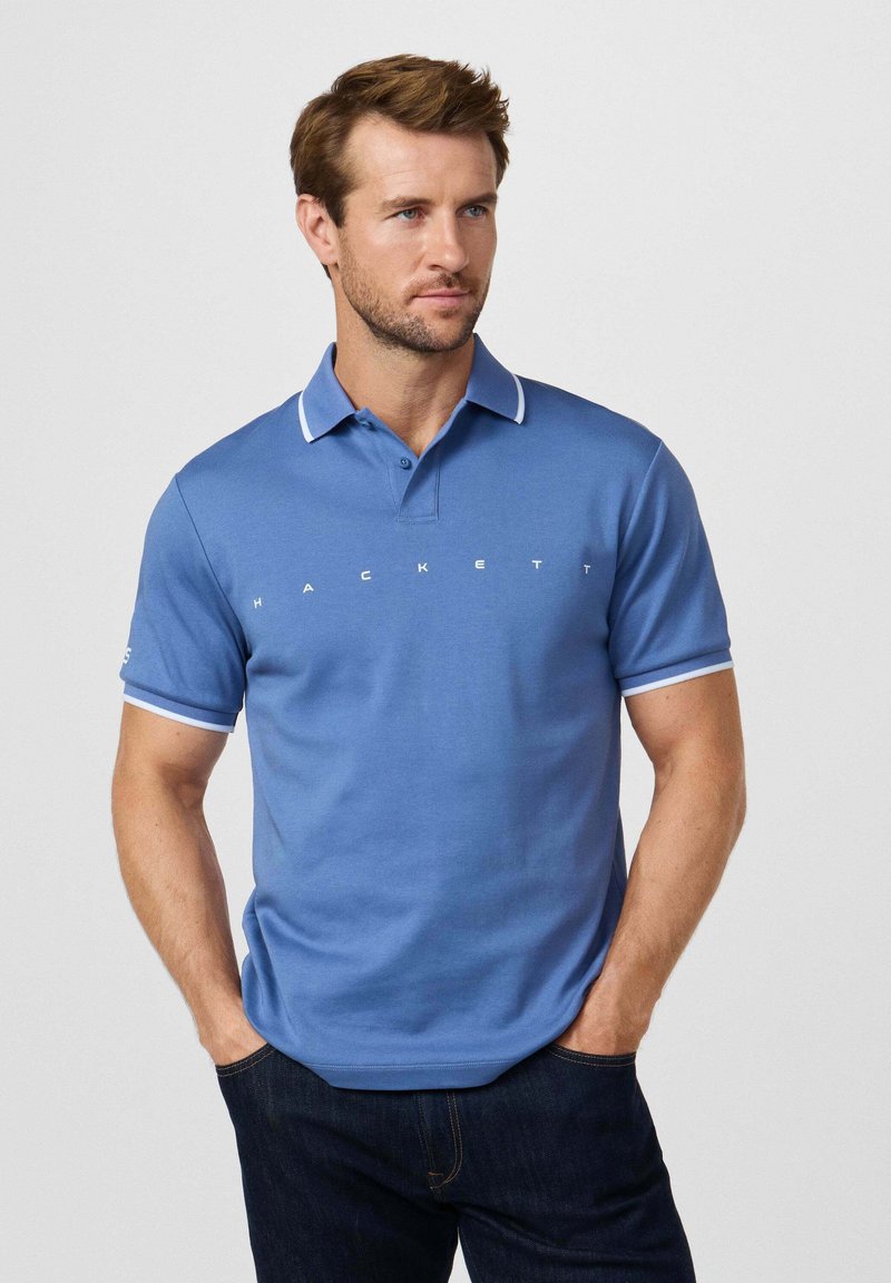 Polo azul claro con detalles blancos en el cuello y las mangas, con un logo "HACKETT" blanco centrado, hecho de tela suave.