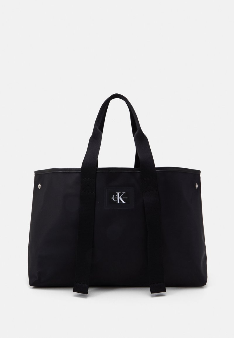 Black tote bag with a rectangular shape, made of durable material, featuring two sturdy handles, a logo patch, and silver hardware accents.