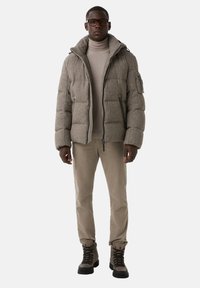 Veste grise matelassée avec col haut et poches zippées, associée à un col roulé beige, un pantalon beige et des bottes à lacets marron.