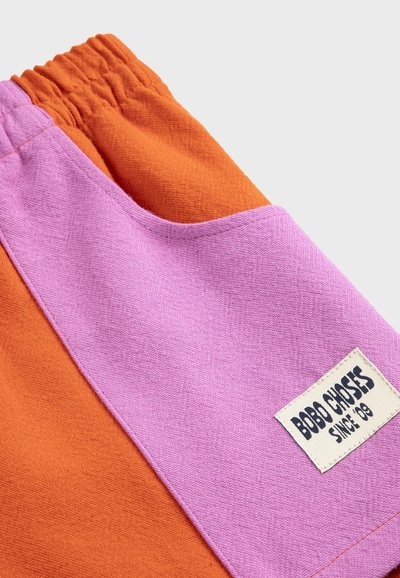Orangene Hose mit einer rosa aufgesetzten Tasche, die ein weißes "Bobo Choses Since '09"-Etikett trägt, wobei die Stoffstruktur und der elastische Bund sichtbar sind.