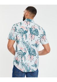 Chemise à manches courtes avec un fond bleu clair, présentant des motifs vibrants de feuilles et de fleurs tropicales en vert et rose. Coupe ajustée avec un ourlet arrondi.