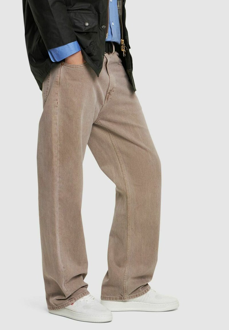 Jeans marrone chiaro a gamba larga realizzati in tessuto robusto, caratterizzati da una texture sottile e tasche frontali, abbinati a sneakers bianche e giacca nera.