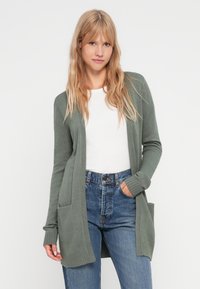Cardigan lung, cu deschidere frontală, din material tricotat moale, verde, cu două buzunare în față și manșete cu dungi, purtat peste o cămașă albă și blugi albaștri.