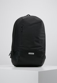 Zaino nero con una parte superiore in sintetico liscio e una base in pelle testurizzata. Presenta una tasca anteriore con zip, un'etichetta con logo e spallacci regolabili.