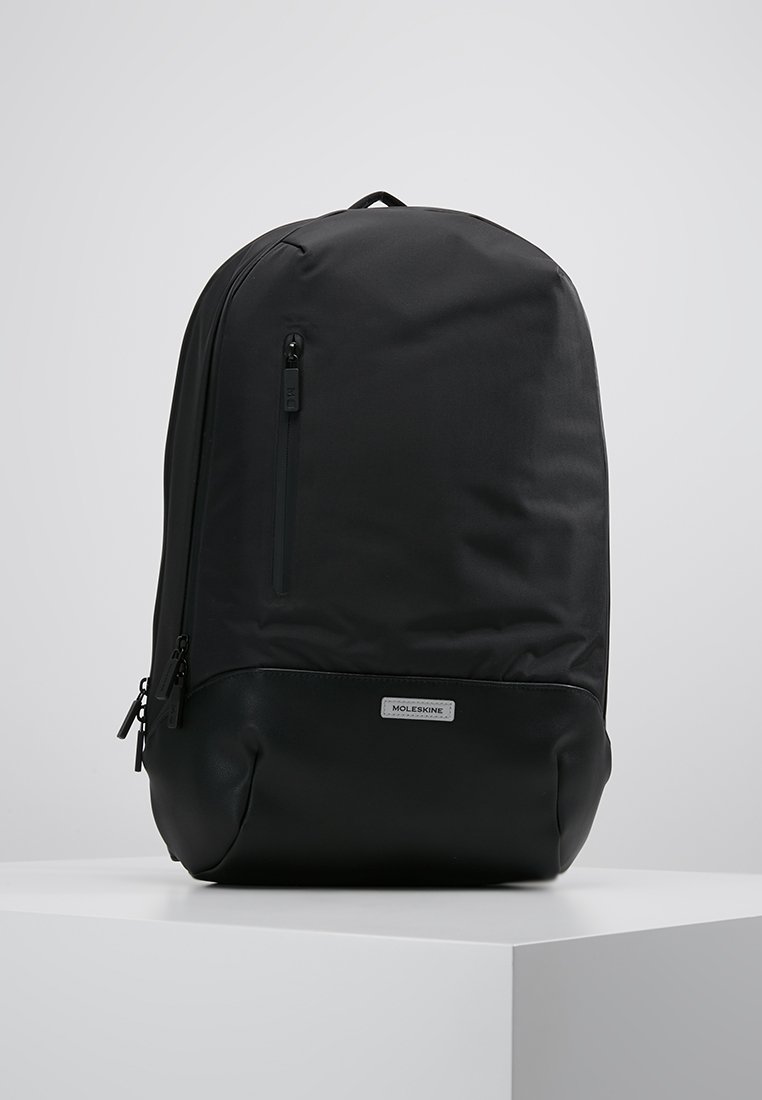 Zaino nero con una parte superiore in sintetico liscio e una base in pelle testurizzata. Presenta una tasca anteriore con zip, un'etichetta con logo e spallacci regolabili.