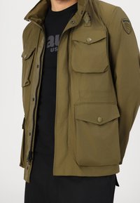 Olivengrøn jakke med lynlås foran, trykknapper og flere store lommer. Lavet af glat stof med en let glans og et mærke på skulderen.
