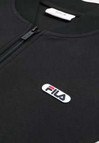 Sort lynlåstrøje med et nærbillede af FILA-logoet i hvid, rød og marineblå på venstre brystområde.