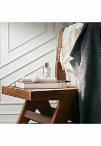 Selkeä lasinen tuoksupullo puupöydällä, jossa on teksturoitu pinta, pinottuja kirjoja, pyöreä valkoinen esine ja pehmeitä kankaita lähellä.