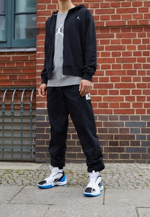Persona de pie en la acera, vistiendo una sudadera con cremallera negra, camiseta gris, pantalones negros con logo y zapatillas blancas, negras y azules en un entorno urbano.