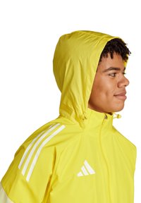 Veste à capuche jaune dotée d'une fermeture éclair, d'un cordon de serrage et d'accents rayés blancs sur les épaules. Tissu léger et texture lisse.