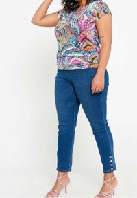 Donna che indossa un top a maniche corte con motivi colorati, jeans skinny blu con bottoni vicino alle caviglie e tacchi a strappo di un leggero viola, in piedi.