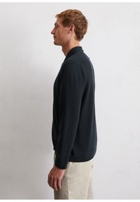 Dark navy knitted polo shirt with long sleeves, a collar, and textured fabric. Worn with light-colored trousers, shown from the side.