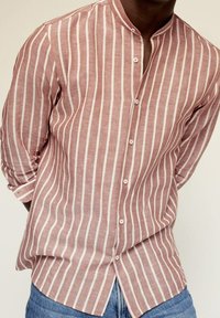 Camisa de lino a rayas en óxido y blanco, con cuello mandarina, cierre frontal de botones y mangas enrolladas. Textura suave con un ajuste relajado.
