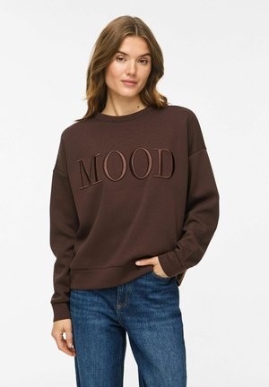 Femme aux cheveux châtain clair portant un sweat-shirt marron foncé avec le texte "MOOD" et un jean bleu, debout avec une main dans sa poche.
