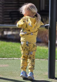 Gul huvtröja och matchande byxor med tecknade djurmönster; barn som har färgglada sneakers; utomhus på gräset med en metallstång.