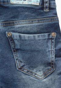 Jeans de mezclilla con un lavado azul medio, que incluyen un bolsillo cosido, herrajes de cobre y una etiqueta con un fondo azul claro.