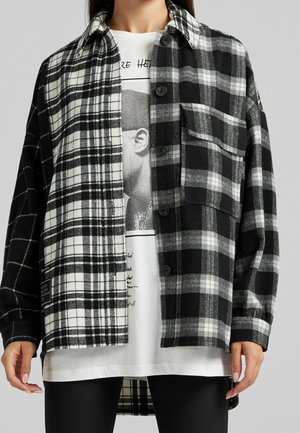 Personne portant une chemise en flanelle à carreaux noir et blanc sur un t-shirt graphique blanc avec un pantalon noir, debout devant un fond uni.