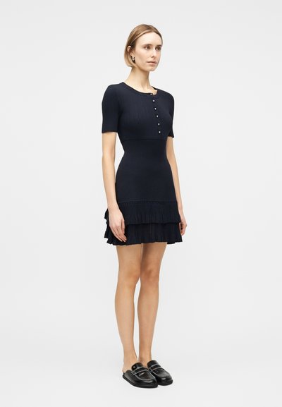 Γυναίκα που στέκεται, φορώντας ένα μπλε ναυτικό φόρεμα με κοντά μανίκια, λεπτομέρειες με κουμπιά στο πάνω μέρος και φούστα με βολάν σε στρώσεις, φορώντας μαύρα λουστρίνια slip-on loafers.