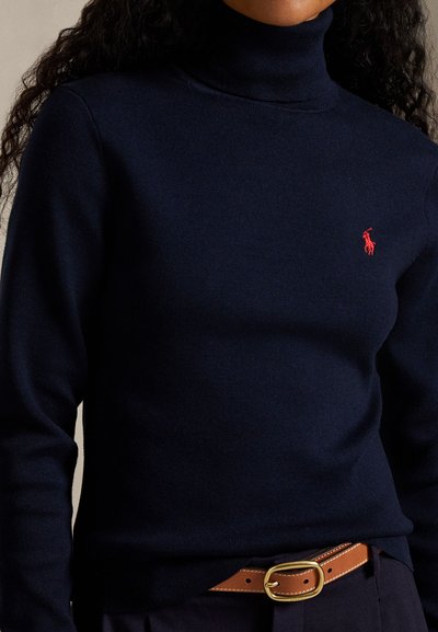 Άτομο που φοράει στενό σκούρο μπλε ζιβάγκο με μικρό κόκκινο κεντημένο λογότυπο στο στήθος και καφέ ζώνη με χρυσή αγκράφα.