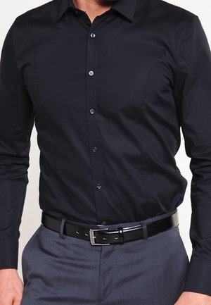 Un homme porte une chemise noire cintrée boutonnée, rentrée dans un pantalon de costume gris avec une ceinture en cuir noire et une boucle argentée.