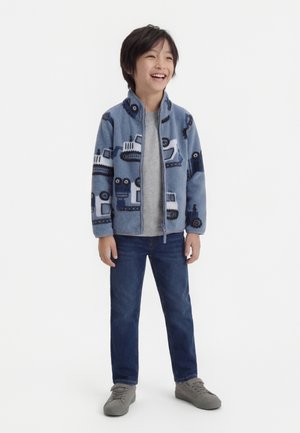 Garçon souriant portant une veste polaire zippée bleue avec des motifs de véhicules de construction, un t-shirt gris, un jean foncé et des baskets grises, debout devant un fond blanc.