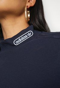 Navy-blaues, geripptes Oberteil mit einem weißen Adidas-Logo am Kragen und hohem Halsausschnitt, ergänzt durch große goldene Creolen.