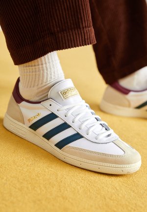 Hvid Adidas Spezial sneaker med marineblå striber, beige ruskindstpe og bordeaux hæl båret med hvide ribstrikkede sokker og brune kordfløjlsbukser på gul gulv.