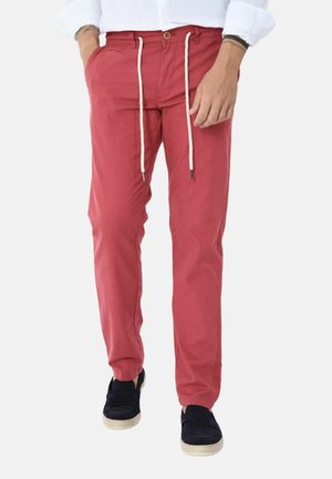 Uomo che indossa pantaloni casual rossi con coulisse bianche, camicia bianca e scarpe slip-on nere su uno sfondo semplice.