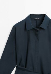 Chemise bleu marine foncé à manches longues avec col pointu et boutons cachés, présentée à plat sur un fond blanc.