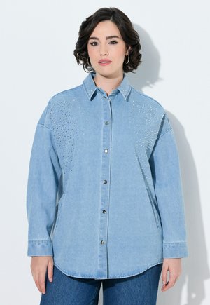 Kvinde med kort mørkt hår iført lyseblå denimskjorte med dekorative nitter og blå jeans mod en ensfarvet hvid baggrund.