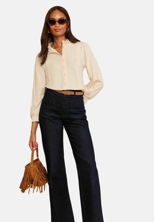 Vrouw met zonnebril, crèmekleurige kanten blouse, donkere broek met hoge taille, houdt een bruine franjehandtas vast met één hand op haar heup.
