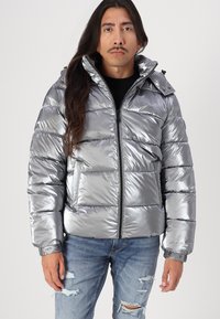 Giacca a vento argentata con finitura metallizzata lucida, dotata di colletto alto, chiusura con zip e tasche frontali. Indossata con jeans in denim strappati.