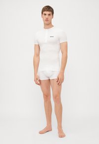 Camicia bianca a maniche corte con patta anteriore e logo, abbinata a slip bianchi. Il tessuto sembra liscio e aderente.