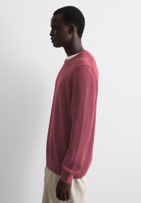 Loose-fitting knit sweater in a textured burgundy shade, featuring a crew neck and long sleeves, paired with light-colored trousers.