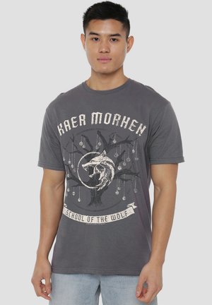 Graues Baumwoll-T-Shirt mit kurzen Ärmeln, gestaltet mit einem Wolfsmotiv, einem Baumgrafik und dem Text "Kaer Morhen" und "Schule des Wolfes".