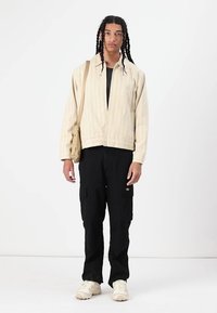 Zip-up cream striped jacket, black cargo pants, and beige sneakers. Model carries a tan tote bag, standing upright against a plain background.