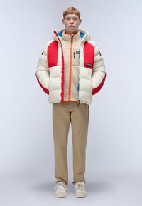 Puffer jacket in cream with red and blue accents, layered over a beige fleece. Paired with beige pants and white sneakers.