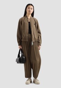 Giacca bomber khaki con orlo elastico, abbinata a pantaloni a gamba larga plissettati. Il modello tiene una borsa nera; delle scarpe beige spesse completano il look.
