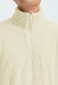 Polarowy sweter w jasnym kremowym kolorze, z wysokim kołnierzem i półzamkiem; tkanina jest miękka i puszysta, o swobodnym fasonie.