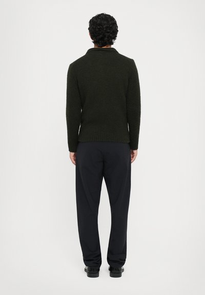 Dunkelgrüner gestrickter Pullover mit Kragen, geripptem Saum und langen Ärmeln, kombiniert mit schwarzen, lässigen Hosen und schwarzen Schuhen.