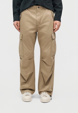 Άτομο που φοράει μπεζ παντελόνι cargo με πλαϊνές τσέπες, μαύρο πουκάμισο και λευκά αθλητικά, που στέκεται μπροστά σε ένα απλό ανοιχτό φόντο.