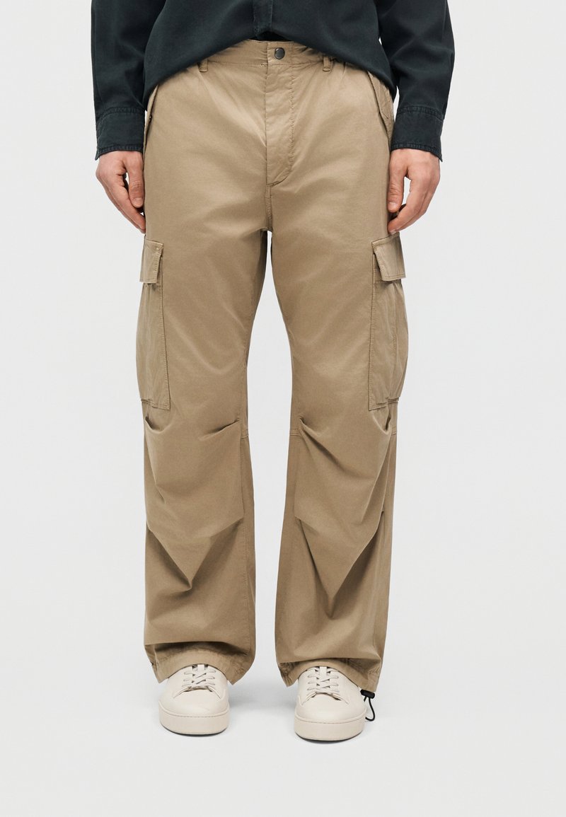 Άτομο που φοράει μπεζ παντελόνι cargo με πλαϊνές τσέπες, μαύρο πουκάμισο και λευκά αθλητικά, που στέκεται μπροστά σε ένα απλό ανοιχτό φόντο.